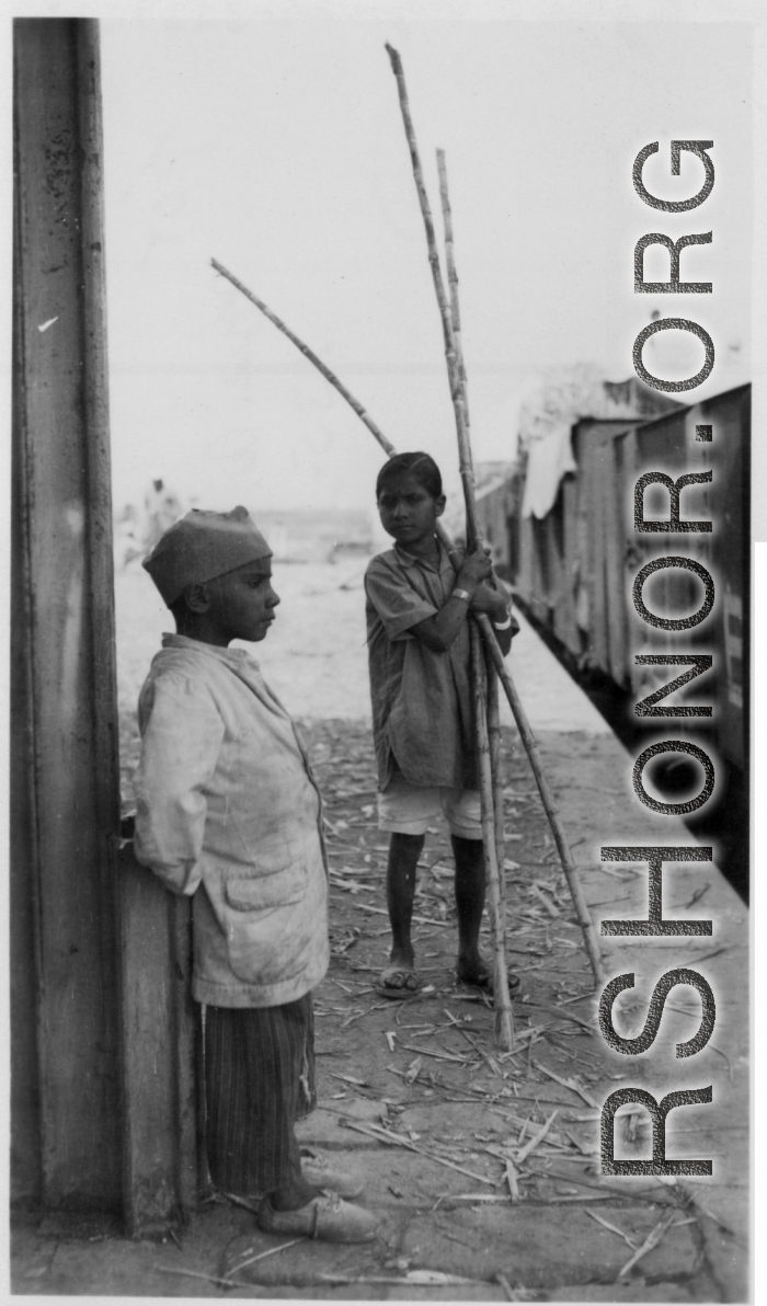 Two boys hang out at train station, one with stalks of sugarcane in hand.  Scenes in India witnessed by American GIs during WWII. For many Americans of that era, with their limited experience traveling, the everyday sights and sounds overseas were new, intriguing, and photo worthy.