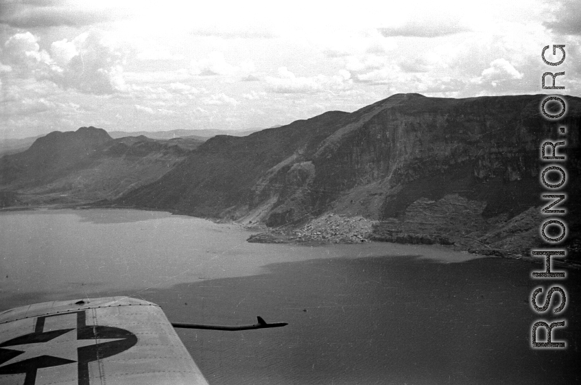 View over wing of Vultee SNV-2 (a variant of the BT-13) trainer piloted by Charles A. Breingan flys over Dianchi Lake (滇池) outside of Kunming, China, during WWII. Small boats dot the water surface, and a large landslide can be seen in the distance. 