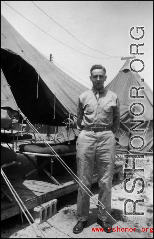 An American stands next to a tent with bed cots, in the CBI, during WWII.