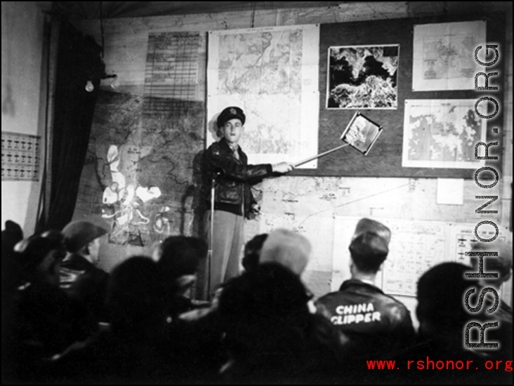 A pre-flight briefing for American flyers in China during WWII.. The maps on the wall behind the speaker clearly show Hong Kong harbor, which the Japanese controlled.