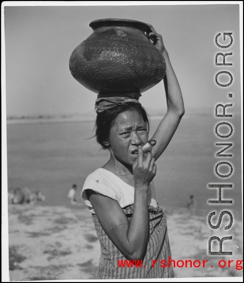 A woman carries a ceramic pot on her head, while smoking an enormous rolled cigarette. In the CBI in WWII.