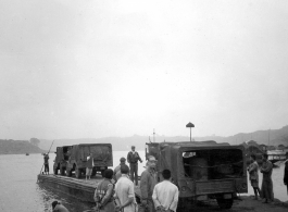 "Vehicles bound for Chengdu and the 20th bomber command are loaded on barges. As a steamboat (behind man directing) waits to ferry the barge across. All necessary gasoline was carried along for the journey. Several 55 gallon drums are visible in the vehicle. Poles were used to move the barges out of shallow water."