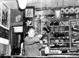 Man in the "Zhang family" hat shop does something to a hanging hat. During WWII. In SW China.