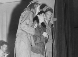 Ladies sing during USO performance in the CBI during WWII. GI plays sax by stage prop labeled for the 748th Railway Operating Battalion.