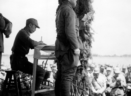 Gen. Wei Lihuang (卫立煌) gives speech to assembled troops, and another person makes notations on paper, on stage during rally. Gen. Du is behind  him on stage.