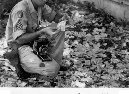 GI examines discarded "invasion currency" on a Rangoon, Burma, street near Bank of India, where it had been dumped during evacuation. Spring 1945.