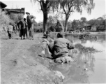 Tall American photographer lugs camera and tripod through village in Yunnan, China, during WWII, while people go about daily life.