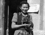 Alice Chong outside the HQ building at Kunming in 1944, holding the Dachshund puppy that her roommate Eloise Witwer gave to General Chennault.  Chennault named the dog “Little Joe” and then changed its name to “Joe-Dog” after it grew up.  The name, of course, was a way of getting back at his nemesis, General Joseph Stilwell because, as Chennault would often say, “Both were low-down, sons of bitches”. . .  Note the notice for the upcoming “Rice Bowl” football game pinned to the door frame too.  This is likel