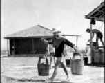A Chinese staff person at an American base in China during WWII carries buckets on a shoulder pole.
